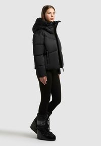Kvinde iført en sort pufferjakke med hætte, sorte leggings og sorte isolerede vinterstøvler, der står mod en simpel lys baggrund.