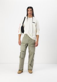 Langærmet cremefarvet skjorte med sort logo, parret med lysegrønne cargo-bukser med flere lommer og brune sko. Sort crossbody taske.