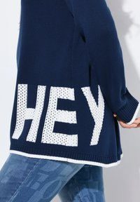 Pull en maille tricoté bleu marine avec un logo blanc "HEY", doté d'un ourlet et de poignets côtelés blancs contrastants, exhibant une texture de tissage ouvert.