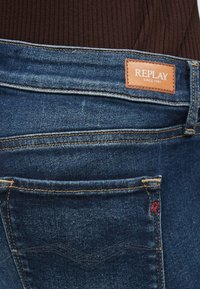 Σκούρο μπλε τζιν με ομαλή υφή, που διαθέτει καφέ δερμάτινο λογότυπο και κόκκινη ραφή στο πίσω τσέπη.