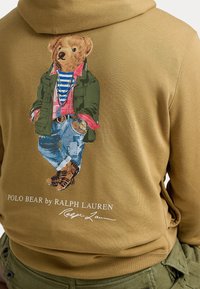 Felpa marrone con un grafico colorato di un orso in abbigliamento casual, con magliette a strati e accessori, e testo del logo sottostante.