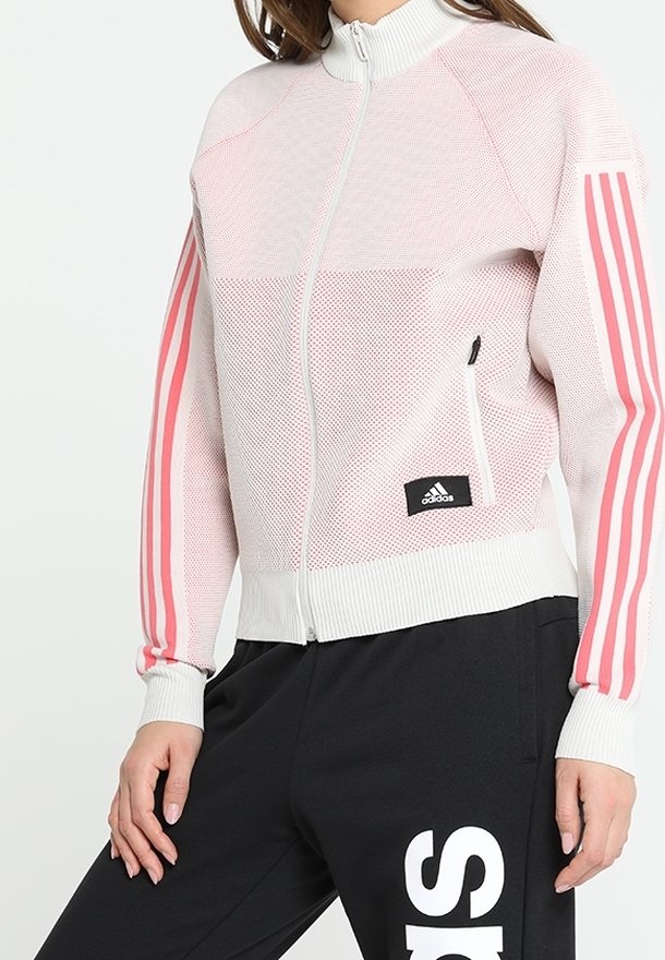 Chaqueta de malla rosa claro con cremallera, que presenta cuellos y puños acanalados, con tres franjas rosas en cada manga y un pequeño parche de logo negro.