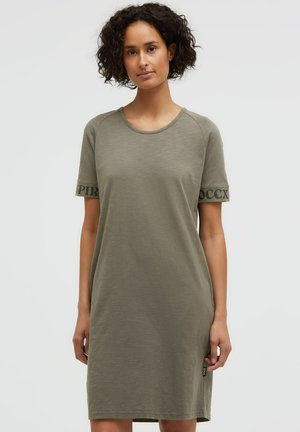 Woman with curly hair wearing a simple olive green short-sleeve dress with text on sleeve edges, standing against a light background.