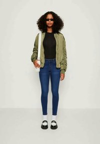 Casaco bombazine verde-oliva com punhos elásticos, blusa preta ajustada, jeans skinny azuis, meias brancas e sapatos pretos de plataforma. Saco tiracolo simples.