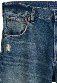 Vaqueros de mezclilla azul con un diseño clásico de cinco bolsillos, áreas desgastadas, herrajes en tono plateado y detalles de costura en contraste.
