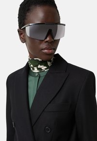 Black oversized sunglasses with gray lenses, worn with a green shirt and a patterned scarf. The blazer features a double-breasted design.