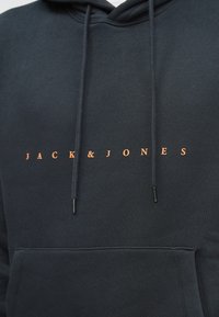 Melnā kapuce ar oranžu "JACK & JONES" tekstu centrā virs priekšējās kabatas un diviem karājošiem auklām.