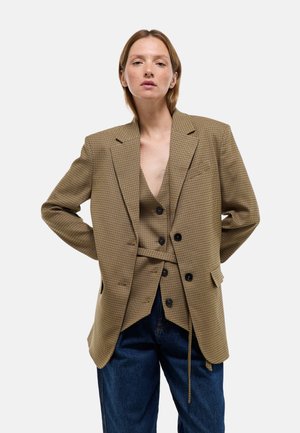 Femme portant un blazer marron à carreaux oversize et un gilet assorti avec un jean bleu taille haute, debout les mains dans le dos.