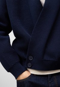 Marinblå stickad kofta med en strukturerad yta, med en V-hals, två svarta knappar och en krämfärgad ribbad nederkant, lager-på-lager över mörk denim.