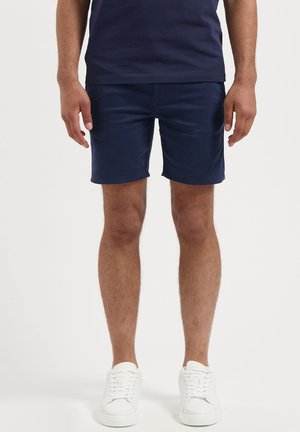 Man wearing navy blue shorts, navy shirt, and white sneakers standing against a plain background.