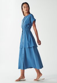 Modra, večstopenjska obleka s kratkimi rokavi in gumbnim zapiranjem spredaj, ki ima elastičen pas in lahkotno krilo do gležnjev.