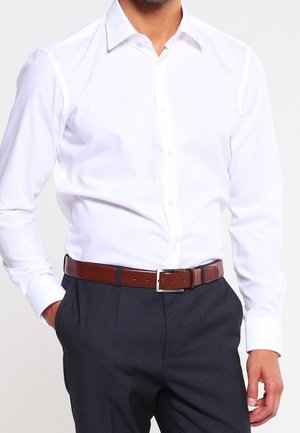Mann trägt ein weißes Hemd mit Knöpfen, dunkle Hosen mit Falten und einen braunen Ledergürtel mit silberner Schnalle. Hände in den Taschen.