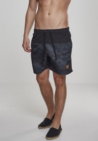 Shorts de bain noirs et camouflage sombre avec une taille élastique et un cordon de serrage, présentant un patch logo beige sur le côté. Chaussures décontractées à enfiler.