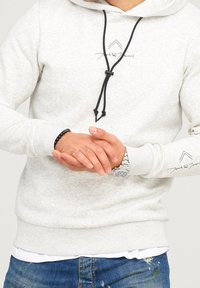 Mann trägt einen hellgrauen Hoodie mit schwarzen Kordeln, Logos auf der Brust und dem Ärmel, blaue Jeans, eine Uhr und ein schwarzes Perlenarmband, Hände gefaltet.