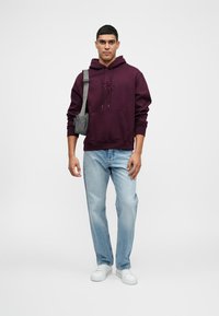 Hanorac colorat căprui cu buzunar kangaroo și logo, blugi albaștri deschiși și adidași albi. O geantă gri este purtată pe umăr.