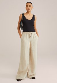 Czarny top na ramiączkach z koronkowym wykończeniem, luźne beżowe lniane spodnie z trokiem w pasie oraz czarne, długie, teksturowane buty na szpilkach. Modelka trzyma czarną torbę.