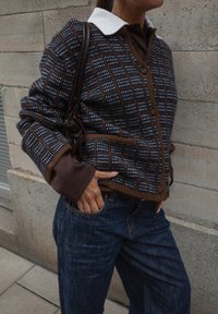 Hnedo-modrý kardigán s vzorom a bielym golierom, prehodený cez hnedú košelu a spárovaný s tmavými riflami, doplnený bočnou taškou.