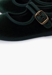 Ballerine in velluto verde scuro con punta rotonda, caratterizzate da una fascia decorativa con una fibbia in bronzo e cuciture a contrasto lungo i bordi.