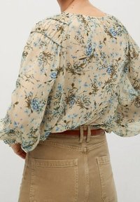 Sheer cream blouse with blue floral pattern, elasticized cuffs, and pleated texture. Worn with light beige high-waisted pants.