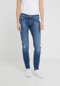 Modré denimové džíny s obtáhnutým střihem, zdobené distressed akcenty a mírně vybledlým povrchem. Nosič má na sobě bílé tenisky.