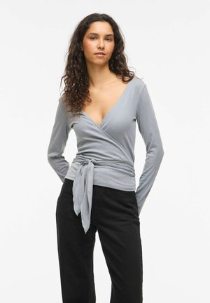 Žena s kučeravými vlasmi, oblečená v svetlosivé zavinovacie dlhé tričko zviazané v páse a čierne nohavice, stojaca na bielom pozadí.