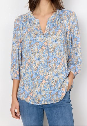 Kvinde iført en bluse med blå, beige og pink blomstermønster med trekvartærmer og blå jeans, den ene hånd i lommen.