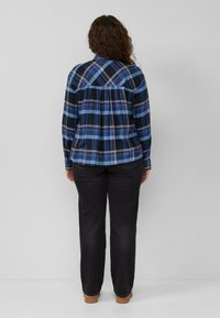 Woman with curly hair wearing a blue plaid shirt, black jeans, and brown shoes, standing facing a plain gray wall.
