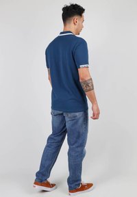 Polo-shirt marine avec col blanc, manches courtes, et logo "OXBOW" sur le poignet. Associé à un jean bleu et des baskets marron. Vue de dos.