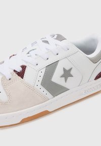 Adidași albi cu accente din piele gri, panou din suede bej, detaliu textil vișiniu și talpă din cauciuc natural. Sunt echipați cu șireturi albe și logo-ul în formă de stea.