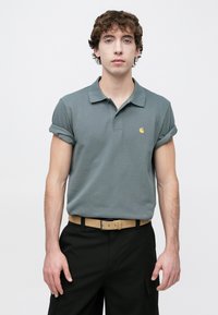 Γκρι κοντομάνικο πόλο πουκάμισο με γυρισμένα μανίκια και μικρό κίτρινο λογότυπο, συνδυασμένο με μαύρο παντελόνι και ταμπά ζώνη.