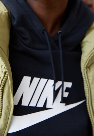 Close-up of person wearing a navy blue Nike hoodie with white logo, partially covered by a light green jacket with visible zipper.