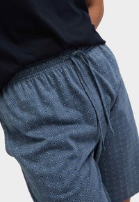 Acercamiento a unos pantalones cortos azules con un estampado de pequeñas formas florales y cuadradas en blanco y azul marino, llevados con una blusa negra. Se ve el cordón en la cinta de la cintura.