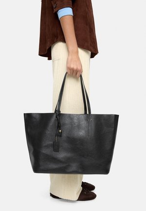 Czarna skórzana torba typu tote z długimi uchwytami, gładką fakturą i małym elementem w postaci metki. Noszona z kremowymi spodniami w prążki i brązową zamszową bluzką.