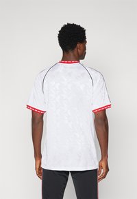 Witte sporttrui met een geometrisch patroon, rode accenten op de kraag en manchetten, zwarte piping langs de naden.
