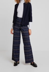 Marineblauwe cardigan met korte mouwen over een grijze coltrui. Hoogwaardige, wijde geruite broek in marineblauw met crèmekleurige en bruine strepen. Zwarte loafers.