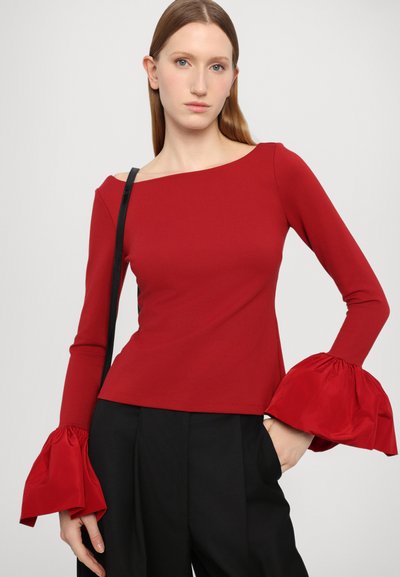 Red fitted top with an off-shoulder neckline and long sleeves featuring voluminous, ruffled cuffs. Paired with black trousers.