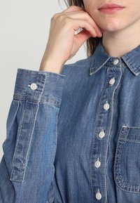 Džínová košile s límečkem, světle modré barvy, s plnou légou na knoflíky, jednou náprsní kapsou a detaily na knoflíkové manžetě. Měkká textura.