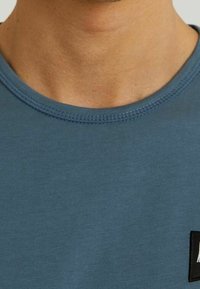 Detailní záběr osoby v modrém tričku s kulatým výstřihem, na kterém jsou viditelné stehy a malá černá obdélníková etiketa na hrudi.