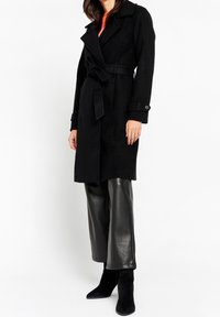 Czarny płaszcz z mieszanki wełny z paskiem w talii, kołnierzem z klapami i zapinanymi na guziki mankietami. Noszony na czarnych szerokich spodniach ze skóry i botkach na obcasie.