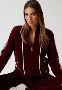 Bordeaux velours zip-up jack met witte strepen op de mouwen en voorkant, met een hoge kraag en een gladde textuur. Maat en pasvorm niet gespecificeerd.