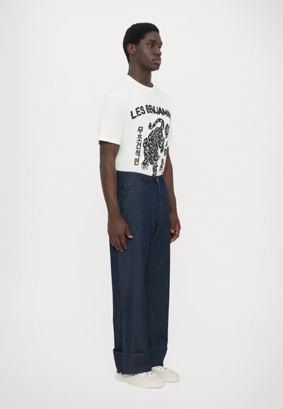 Λευκό κοντομάνικο μπλουζάκι με μαύρη γραφική εκτύπωση, συνδυασμένο με φαρδιά σκούρα μπλε τζιν και λευκά αθλητικά παπούτσια.