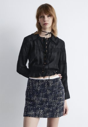 Schwarze Bluse mit Knöpfen, Spitzenakzenten und Rüschensaum, kombiniert mit einem strukturierten Marineblauen Rock mit einem weiß-beigen Karomuster.