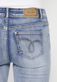 Jean bleu clair avec une patch en cuir, des coutures décoratives sur la poche arrière et une coupe slim.