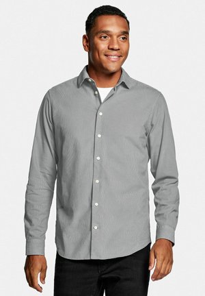 Man in een lichtgrijs overhemd met lange mouwen over een wit ondershirt, glimlachend en naar rechts kijkend tegen een witte achtergrond.