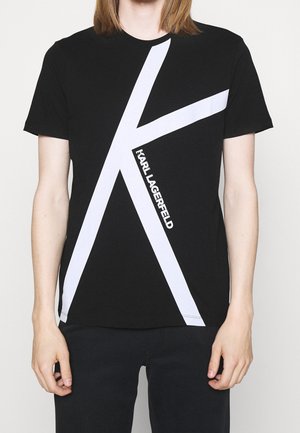 Person trägt ein schwarzes T-Shirt mit großem weißem "K"-Design und vertikal gedrucktem "KARL LAGERFELD"-Schriftzug auf der Vorderseite.