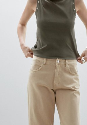 Osoba ubrana w oliwkowozielony top bez rękawów z guzikami oraz beżowe spodnie z wysokim stanem, poprawiająca dół topu.