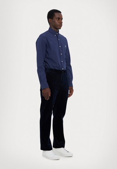 Mørkeblå skjorte med hvite prikker og knappelukking, kombinert med mørke fløyelsbukser og hvite joggesko, som skaper en strukturert silhuett.