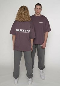 Oversized, lilla t-shirts med korte ærmer og teksten "MULTIPLY APPAREL CLUB" i hvidt på ryggen. Kombineret med grå joggers og hvide sneakers.