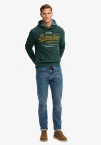 Zielona bluza z kapturem z przednią kieszenią, z żółtym logo. Połączona z niebieskimi jeansami i brązowymi sznurowanymi butami. Swobodny krój.