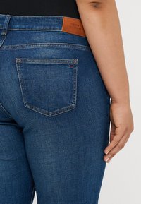 Tumed sinine teksariie, millel on sile tekstuur, tagakotis ja pruun nahklabel, millel on peale trükitud "TOMMY HILFIGER."